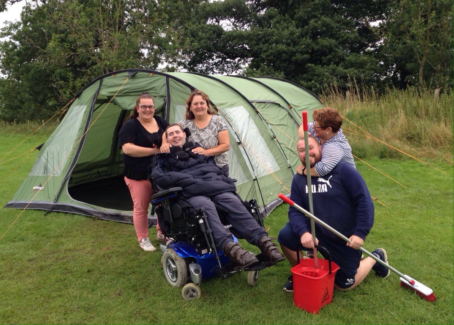 Male with disabilities in electric wheelchair with friends and carers infront of a tent in a field